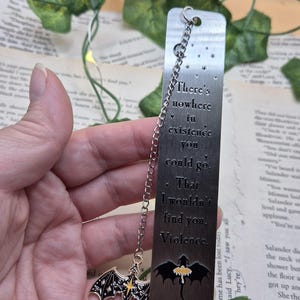 May include: A silver metal bookmark with a chain and two charms. The bookmark has the quote "There's nowhere in existence you could go. That I wouldn't find you. Violence." A black dragon charm hangs from the chain.
