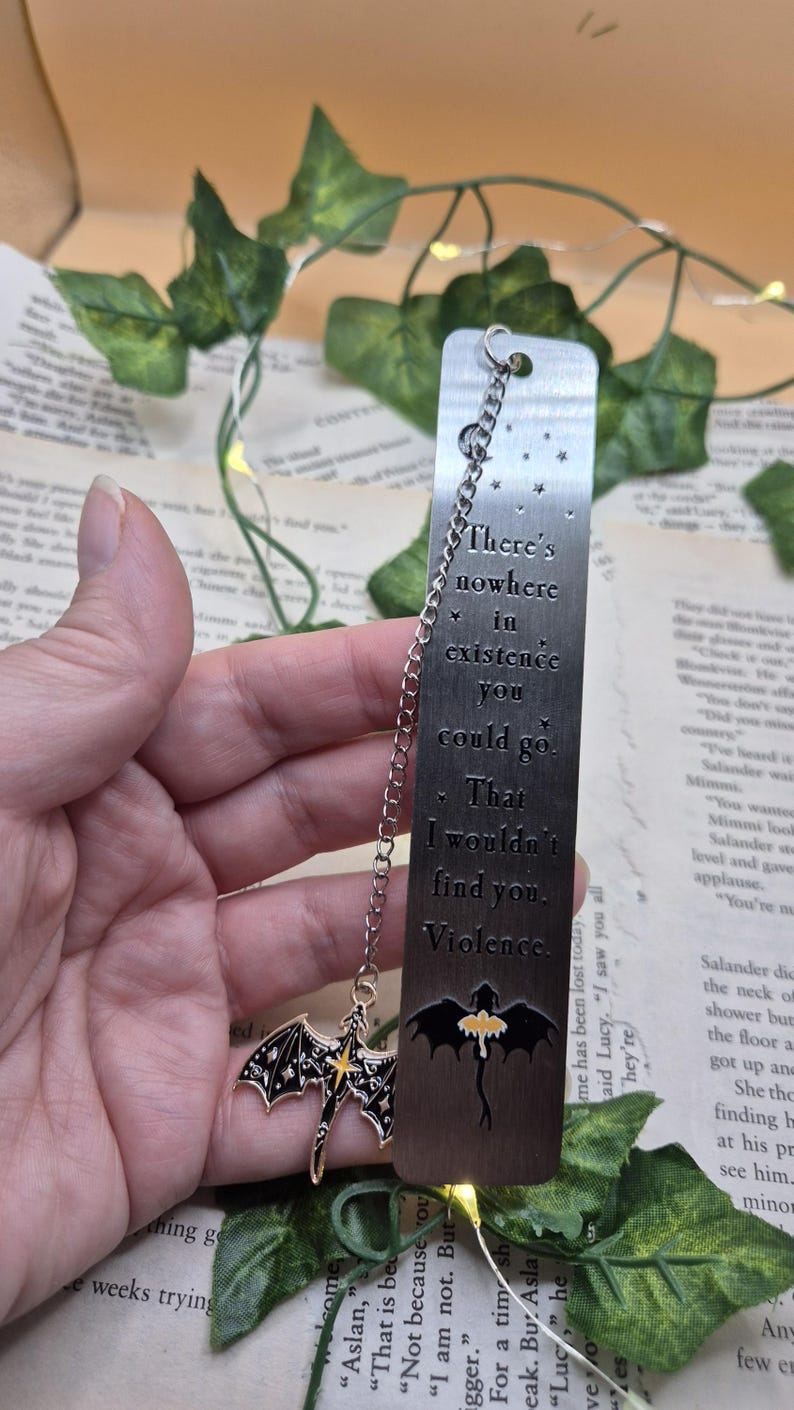 May include: A silver metal bookmark with a chain and two charms. The bookmark has the quote "There's nowhere in existence you could go. That I wouldn't find you. Violence." A black dragon charm hangs from the chain.