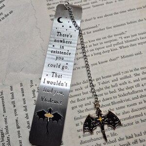 May include: A metal bookmark with a chain and a bat charm. The bookmark has etched text: "There's nowhere in existence you could go. That I wouldn't find you, Violence." The bat charm has a yellow cross.