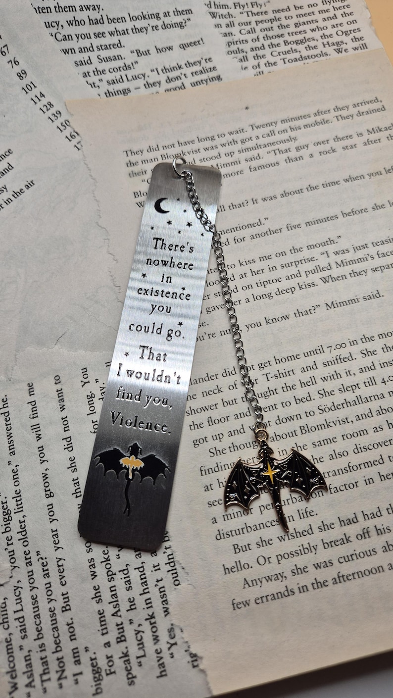 May include: A metal bookmark with a chain and a bat charm. The bookmark has etched text: "There's nowhere in existence you could go. That I wouldn't find you, Violence." The bat charm has a yellow cross.