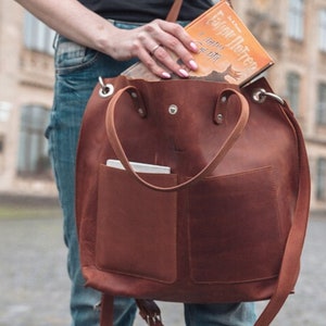Puede incluir: Un bolso tote de cuero marr&oacute;n con un bolsillo grande en la parte delantera. El bolso tiene una correa larga y dos asas m&aacute;s cortas. Un libro titulado "Harry Potter y las Reliquias de la Muerte" es parcialmente visible dentro del bolso.