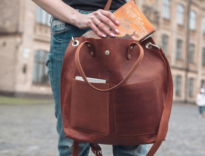 Puede incluir: Un bolso tote de cuero marr&oacute;n con un bolsillo grande en la parte delantera. El bolso tiene una correa larga y dos asas m&aacute;s cortas. Un libro titulado "Harry Potter y las Reliquias de la Muerte" es parcialmente visible dentro del bolso.