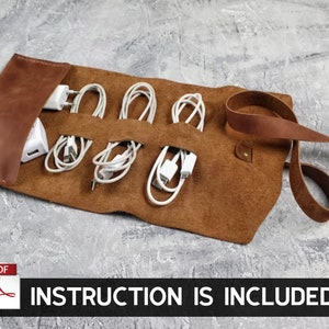 Può includere: Organizzatore di cavi in ​​cuoio marrone con sei fessure per i cavi e un cinturino per fissare il rotolo. L'organizzatore è srotolato e contiene cavi e caricabatterie bianchi all'interno.