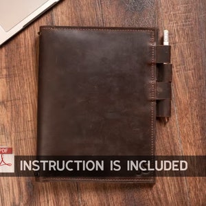 May include: A dark brown leather journal with a pen holder and a pencil. The journal is lying on a wooden surface. The text "INSTRUCTION IS INCLUDED" is visible.