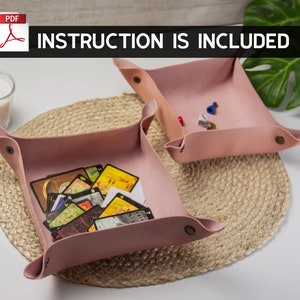 Könnte beinhalten: Zwei rosa Ledertabletts, eines mit einem Kartenspiel und das andere mit Würfeln und einer Spielfigur. Die Tabletts befinden sich auf einer geflochtenen Strohmatte. Der Text "INSTRUCTION IS INCLUDED" befindet sich oben im Bild.