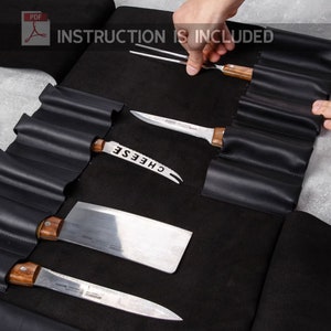 Op de afbeelding: Zwarte leren messenrol met meerdere vakken voor messen en gereedschap. De rol is open en toont een verscheidenheid aan messen, waaronder een kaasmesser, een koksmes en een schilmes. De rol is gelabeld met "INSTRUCTION IS INCLUDED".