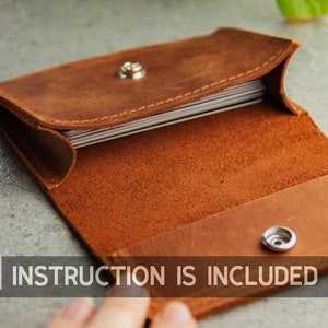 May include: Open, brown leather business card holder with a snap closure. The interior reveals a stack of cards. The text "INSTRUCTION IS INCLUDED" is visible in the lower left corner.