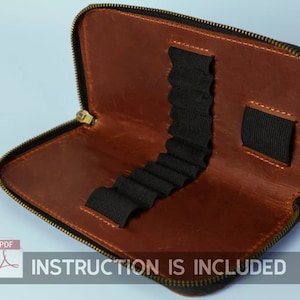 Op de afbeelding: Een bruine leren etui met rits en elastische lussen om potloden vast te houden. De etui is open en het interieur is zichtbaar. De tekst "INSTRUCTION IS INCLUDED" is onderaan de afbeelding gedrukt.