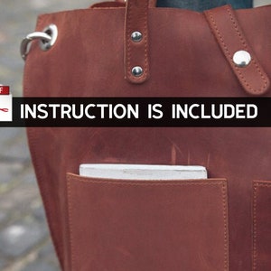 Puede incluir: Un bolso tote de cuero marr&oacute;n con un bolsillo con cremallera y un cierre de solapa. El bolso tiene un papel blanco dentro del bolsillo. El texto "INSTRUCTION IS INCLUDED" est&aacute; impreso en una pancarta negra.