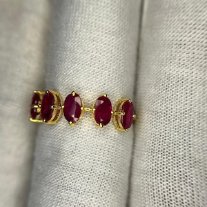 May include: A gold ring with five oval-cut, deep red gemstones. The gemstones are set in individual prongs along the band. The ring is displayed on a white, textured surface.