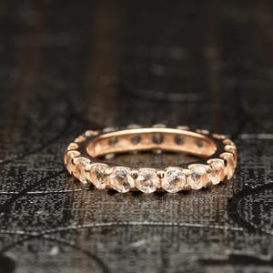 Puede incluir: Un anillo de eternidad de oro rosa con una fila continua de piedras preciosas redondas y claras. El anillo se encuentra sobre un fondo oscuro y texturizado, resaltando el brillo de las piedras.