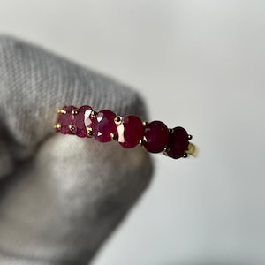 May include: A gold ring with six oval-cut, deep red rubies set in a row. The gemstones are closely aligned, forming a continuous line of colour. The ring has a simple, polished gold band.