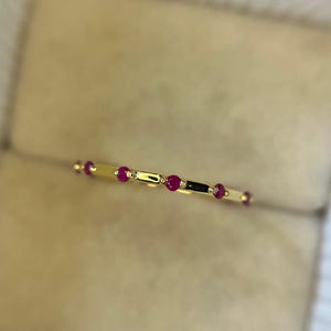 Puede incluir: Un delicado anillo de oro con una serie de pequeñas y vibrantes piedras preciosas rojas. El diseño del anillo incorpora una disposición lineal de las piedras rojas, intercaladas con segmentos dorados. El anillo se presenta sobre un fondo suave y neutro.