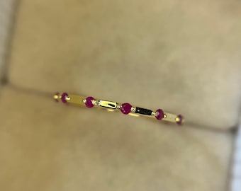 May include: A slender gold ring adorned with a sequence of small, vivid red gemstones. The ring's design includes a linear pattern of the red stones, alternating with gold sections. The ring is displayed against a soft, neutral backdrop.