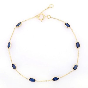 Könnte beinhalten: Ein goldfarbenes Armband mit ovalen, tiefblauen Edelsteinen. Die feine Kette hat gleichmäßig verteilte Steine. Der Verschluss befindet sich oben. Das Armband ist vor weißem Hintergrund abgebildet. Die Steine sind ca. 0,5 cm lang.