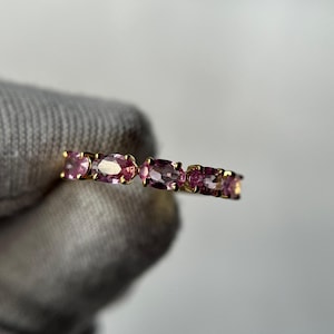 Puede incluir: Un anillo de oro con una serie de piedras preciosas rosas de forma ovalada. Las piedras están engastadas juntas, creando una banda continua de color. El anillo se sostiene sobre un fondo neutro, resaltando el brillo de las piedras.