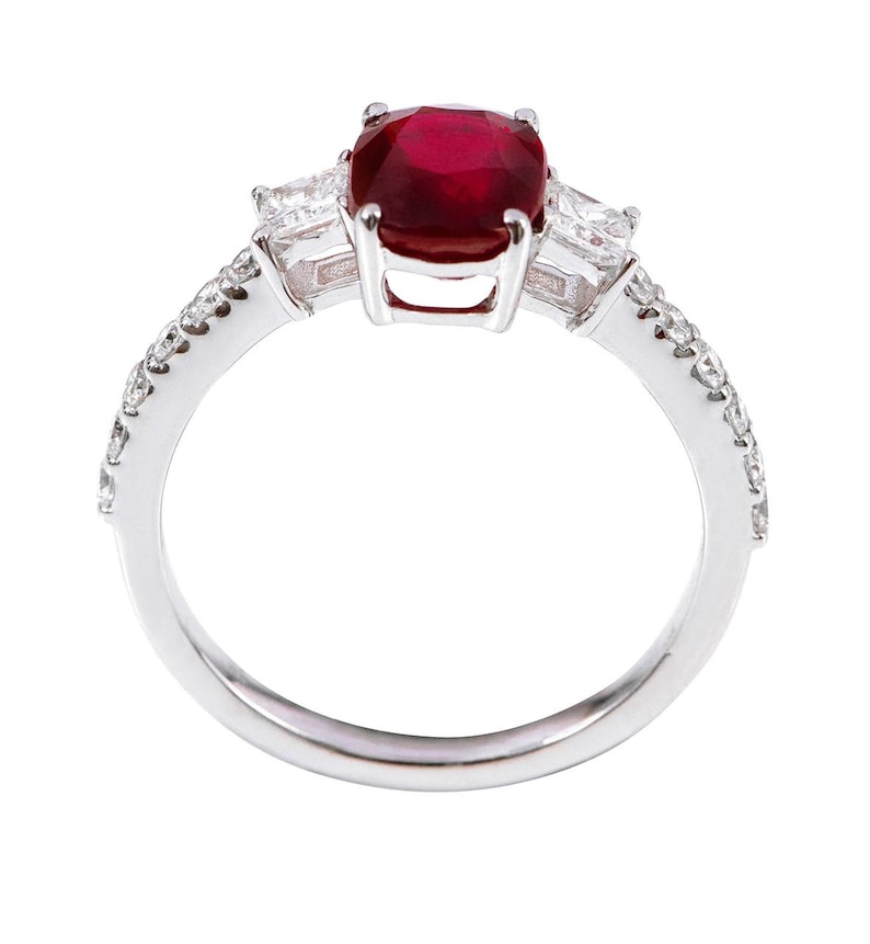 K&ouml;nnte beinhalten: Ein silberner Ring mit einem gro&szlig;en roten Edelstein und zwei kleineren klaren Edelsteinen auf der Ringschiene. Die Ringschiene ist mit kleinen klaren Edelsteinen besetzt.