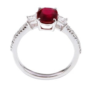 K&ouml;nnte beinhalten: Ein silberner Ring mit einem gro&szlig;en roten Edelstein und zwei kleineren klaren Edelsteinen auf der Ringschiene. Die Ringschiene ist mit kleinen klaren Edelsteinen besetzt.