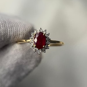 Pode incluir: Um anel de ouro com uma gema vermelha oval, rodeada por um halo de pedras menores e transparentes. A banda do anel é fina e polida, criando um design clássico e elegante. As pedras preciosas brilham contra um fundo neutro.