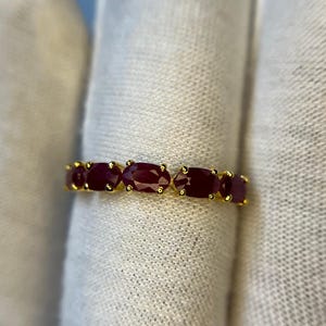 Puede incluir: Un anillo de oro con una serie de piedras preciosas ovaladas de color rojo intenso. Las piedras están engastadas juntas, creando una banda continua de color. La banda del anillo es de color oro brillante, contrastando con las piedras rojas oscuras.