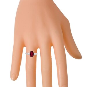 K&ouml;nnte beinhalten: Ein silberner Ring mit einem gro&szlig;en ovalen Rubin, der von kleineren Diamanten umgeben ist. Der Ring wird an einem Modellfinger getragen.