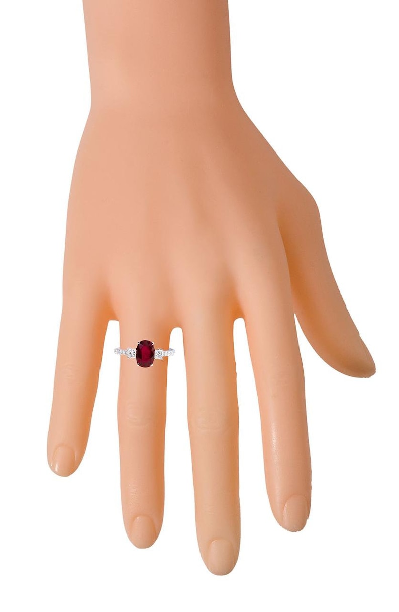 K&ouml;nnte beinhalten: Ein silberner Ring mit einem gro&szlig;en ovalen Rubin, der von kleineren Diamanten umgeben ist. Der Ring wird an einem Modellfinger getragen.