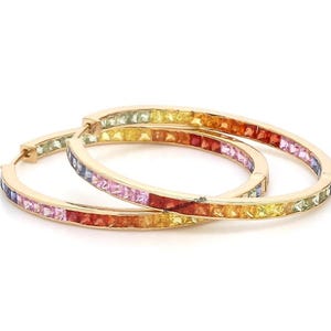 Peut inclure: Une paire de créoles dorées. Chaque boucle d'oreille est ornée d'un arc-en-ciel de pierres précieuses taillées en carré. Les pierres précieuses passent du vert au jaune, à l'orange, au rouge, au rose et au violet. Les boucles d'oreilles sont sur fond blanc.