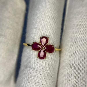 May include: A gold ring featuring a four-petal flower design. Each petal is a pear-shaped, deep red gemstone. The ring is set against a white background.