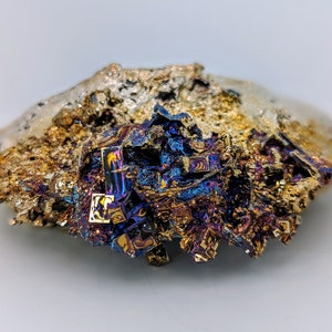 May include: A close-up of a metallic, iridescent mineral specimen with a rough, textured surface. The mineral has a variety of colors, including gold, blue, purple, and black. The specimen is irregularly shaped and appears to be a natural formation.