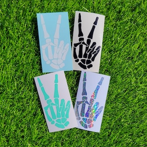 May include: Four peace sign stickers with a skeleton hand design. The stickers are in different colors: white, black, turquoise, and iridescent.