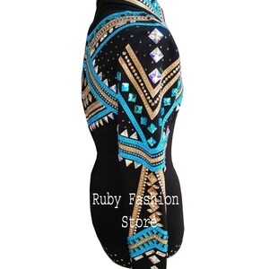 Op de afbeelding: Een zwart shirt met lange mouwen voorzien van ingewikkelde patronen van turquoise, gouden en iriserende kristallen in geometrische vormen over de schouder en mouw. De tekst Ruby Fashion Store is in het wit op de zijkant van het kledingstuk gedrukt.