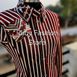 Könnte beinhalten: Ein weiss-rot gestreiftes Hemd mit Kragen und langen Ärmeln. Der Kragen und die Manschetten sind mit roten und klaren Strasssteinen verziert. Der Text "Ruby Fashion Store" ist auf der Vorderseite aufgedruckt.