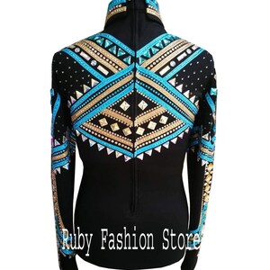 Op de afbeelding: Een zwart shirt met lange mouwen, een hoge kraag en ingewikkelde geometrische patronen in turquoise, goudkleurige en heldere kristallen. Het ontwerp bevat rijen strass-steentjes en studs op de schouders en rug. Tekst: Ruby Fashion Store.