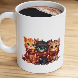 May include: White ceramic mug featuring a design of three kittens wearing scarves. The kittens are orange, black and brown. The mug is filled with coffee.