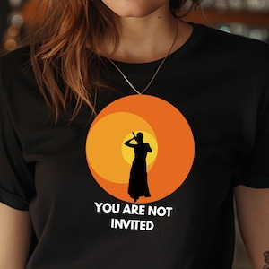Puede incluir: Camiseta negra con un diseño de puesta de sol naranja y la silueta de una persona con un vestido largo. El texto "YOU ARE NOT INVITED" está debajo. Cuello redondo y mangas cortas.