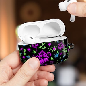 May include: Black AirPod case with a colorful graffiti design featuring a skull, stars, and the word "HA HA". The case is open, revealing white AirPods. A hand holds one AirPod, while the case is held in another hand.