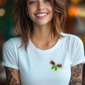 May include: White t-shirt with a green and purple butterfly with gold accents.