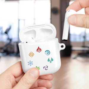 May include: White AirPods case with colorful emblem designs. The case is open, revealing the earbuds inside. One earbud is held separately. The case has a small loop for attaching to a keychain or lanyard. The background is blurred.