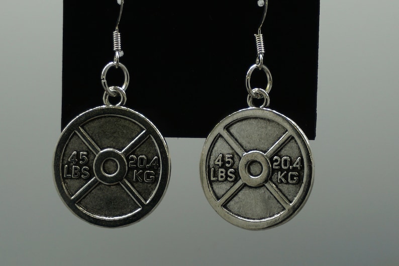 Puede incluir: Un par de pendientes de plata con forma de placa de peso sobre un fondo negro. Las placas son redondas y tienen un dise&ntilde;o de cruz negra con el texto "45 LBS 20.4 KG" en ellas.
