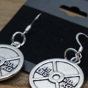 Puede incluir: Pendientes de plata con forma de placa de peso con la inscripci&oacute;n 45 LBS y 20,4 KG. Los pendientes est&aacute;n colgados de una tarjeta de exhibici&oacute;n negra.
