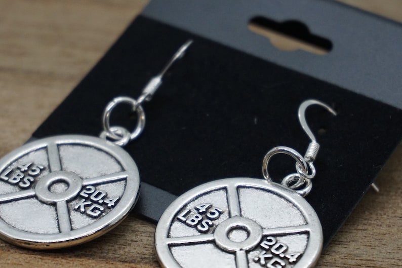 Puede incluir: Pendientes de plata con forma de placa de peso con la inscripci&oacute;n 45 LBS y 20,4 KG. Los pendientes est&aacute;n colgados de una tarjeta de exhibici&oacute;n negra.