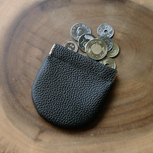 May include: A black leather coin purse with a textured surface, partially open and filled with various coins. The purse is set on a wooden surface, showcasing the contrast between the dark leather and the light wood.