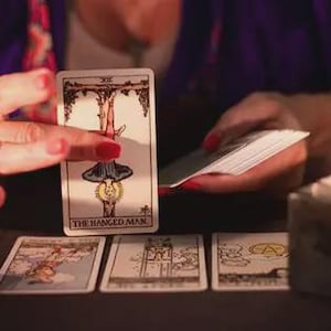 Può includere: Una lettura di tarocchi in corso. La carta dell'Appeso è esposta, con altre carte disposte. Una candela accesa fornisce una luce soffusa. La mano del lettore, adornata da un grande anello, tiene la carta.