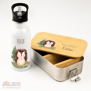 Könnte beinhalten: Eine weiße Wasserflasche und eine Metall-Lunchbox mit Holzdeckel. Beide Artikel zeigen ein Cartoon-Igel-Design mit dem Namen "Felix". Der Lunchbox-Deckel trägt den Text "HIER KRÜMELT Felix".