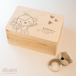 Op de afbeelding: Een lichtgekleurde houten kist met een cartoon aap illustratie en de naam "Emma" gegraveerd op het deksel. De kist wordt vergezeld door een gebreid speelgoed en een houten ring rammelaar. Het deksel bevat de geboortegegevens van de baby.