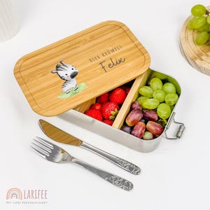 Op de afbeelding: Een bamboe lunchbox met een roestvrijstalen binnenkant, gevuld met aardbeien, druiven en een zebra-illustratie. Het deksel heeft de tekst "Hier Krümelt Felix". Inclusief vork en mes.