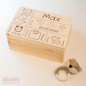 Pode incluir: Caixa de madeira clara com gravuras: uma raposa, o nome "Max" e detalhes de nascimento: 2700g de peso, 51cm de comprimento e a data 24/03/2025. Um chocalho de raposa de malha está ao lado.