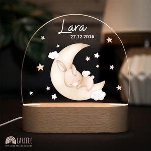 Peut inclure: Veilleuse avec un lapin endormi sur un croissant de lune, entouré d'étoiles et de nuages. Le nom "Lara" et la date "27.12.2016" sont affichés sur l'acrylique transparent. La base est en bois.
