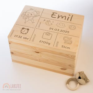 Könnte beinhalten: Eine hellfarbene Holzkiste mit Scharnierdeckel und eingravierten Details. Der Deckel zeigt einen Cartoon-Affen, den Namen "Emil" und Geburtsdaten: Uhrzeit, Datum, Gewicht (2700g) und Größe (51cm). Ein kleines Spielzeug liegt daneben.