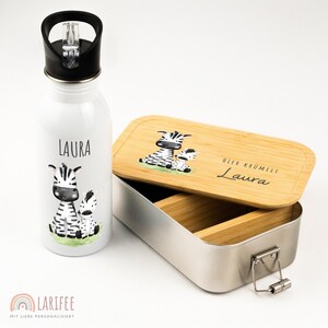 Op de afbeelding: Een witte waterfles en een lunchbox met een zebra thema. De waterfles heeft een zwarte dop en de naam "LAURA" erop gedrukt. De lunchbox heeft een bamboe deksel met de naam "Laura" en de tekst "HIER KRÜMELT" erop.